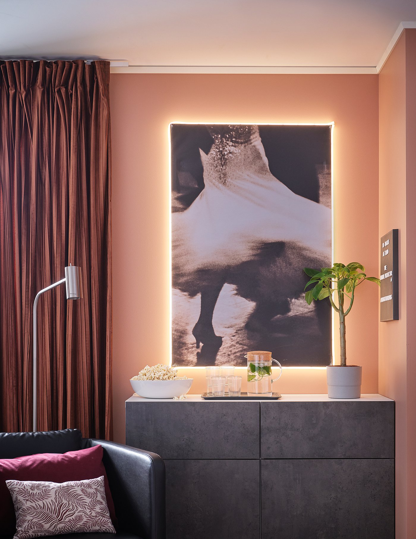 Modern living room with light strips; large abstract photo, dark furniture, and red curtains.