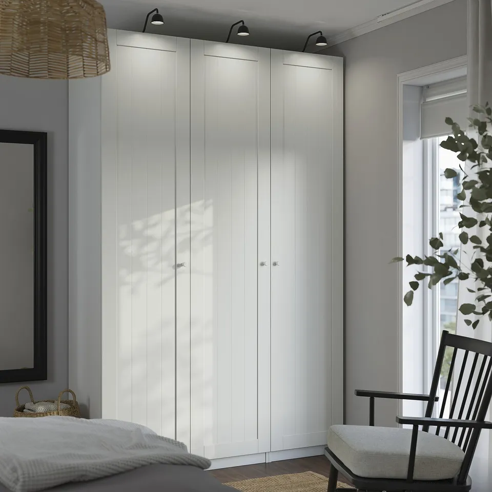 Modern minimalist bedroom with large white PAX wardrobe, tall and spanning wall, beside black framed window and wooden chair.