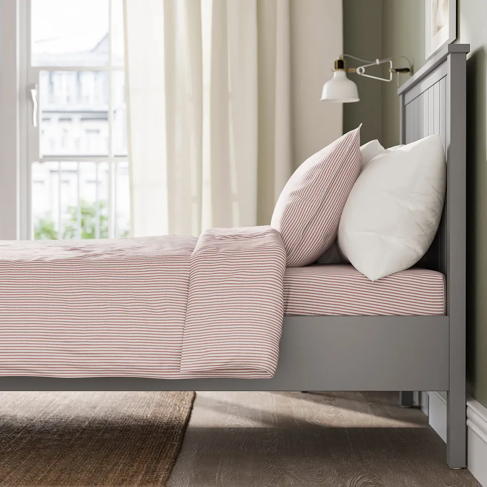 Bed with striped sheets and pillows, wall lamp, large window with curtains.