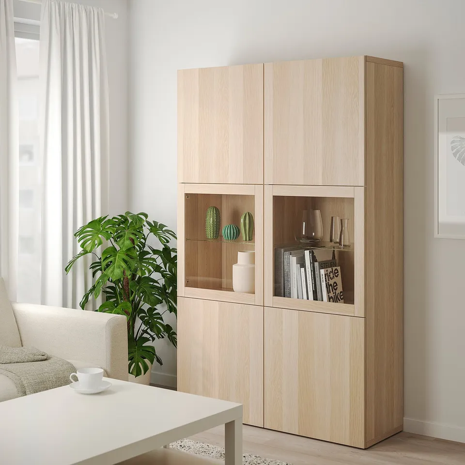 Brown ikea BESTÅ storage unit with glass doors in living room, holding vases and books, beside a white couch.