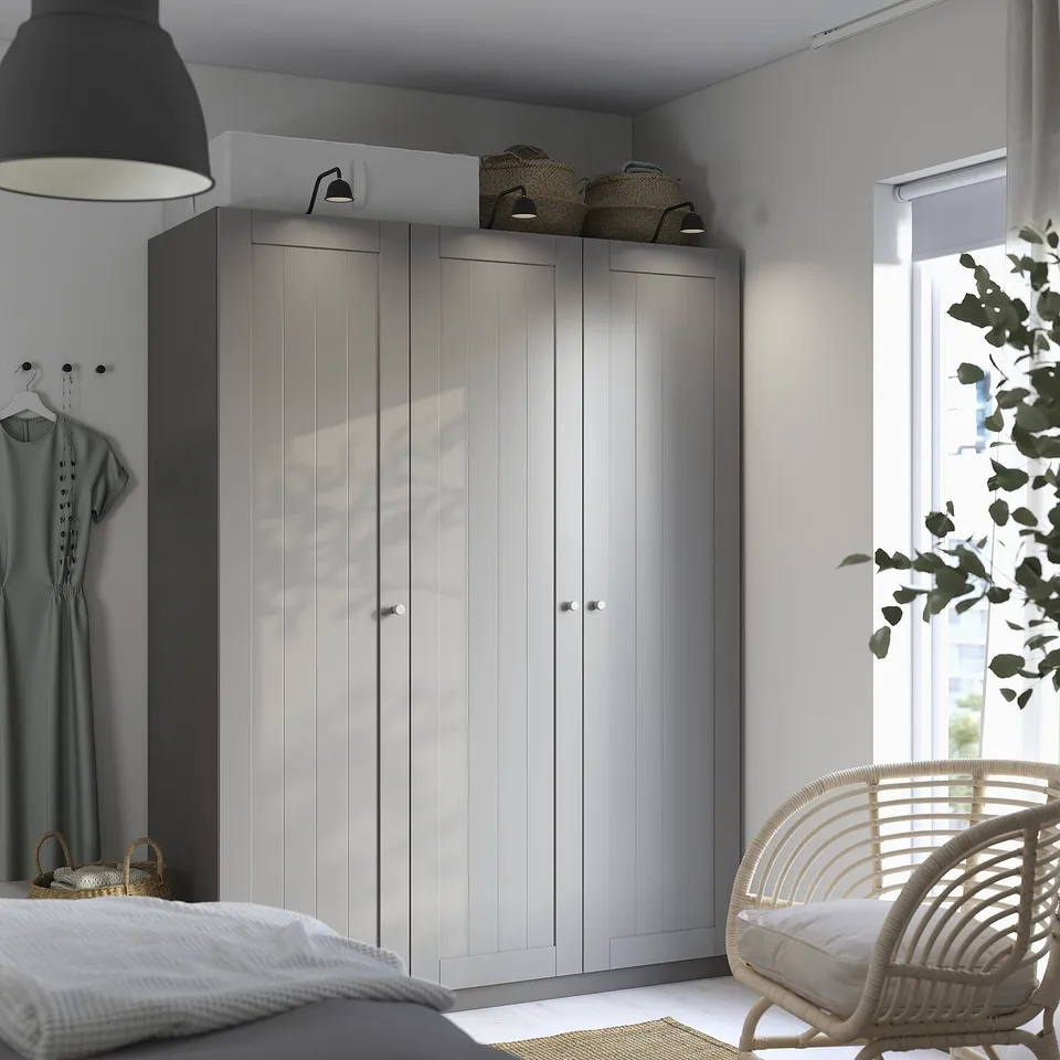Minimalist bedroom with large white PAX wardrobe, wicker chair, and wall lamp. Dress hangs to the left, bed visible at the bottom.