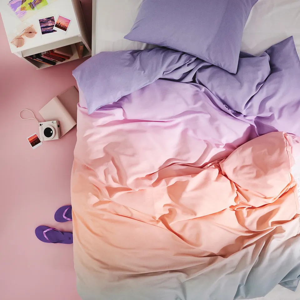 Colourful unmade bed with purple-pink FLÄDERBUSKE duvet, lightweight polyester/viscose, purple slippers, small camera, and photoshelf with sunglasses.
