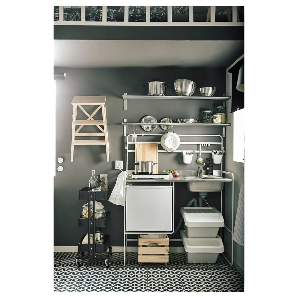 Compact, modern mini-kitchen with stainless steel sink, white fridge, and open shelving. Black and white patterned floor tiles.