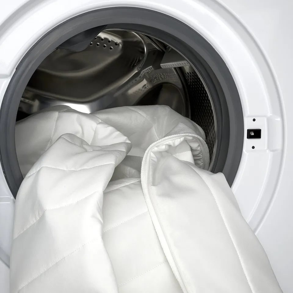 Image depicts a white, padded mattress topper inside a washing machine, emphasising its machine-washable feature for easy cleaning.