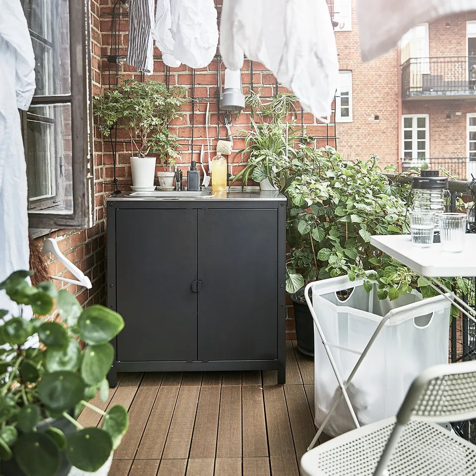 Balcony with black GRILLSKÄR cabinet, plants, hanging laundry, white chairs, brick building.