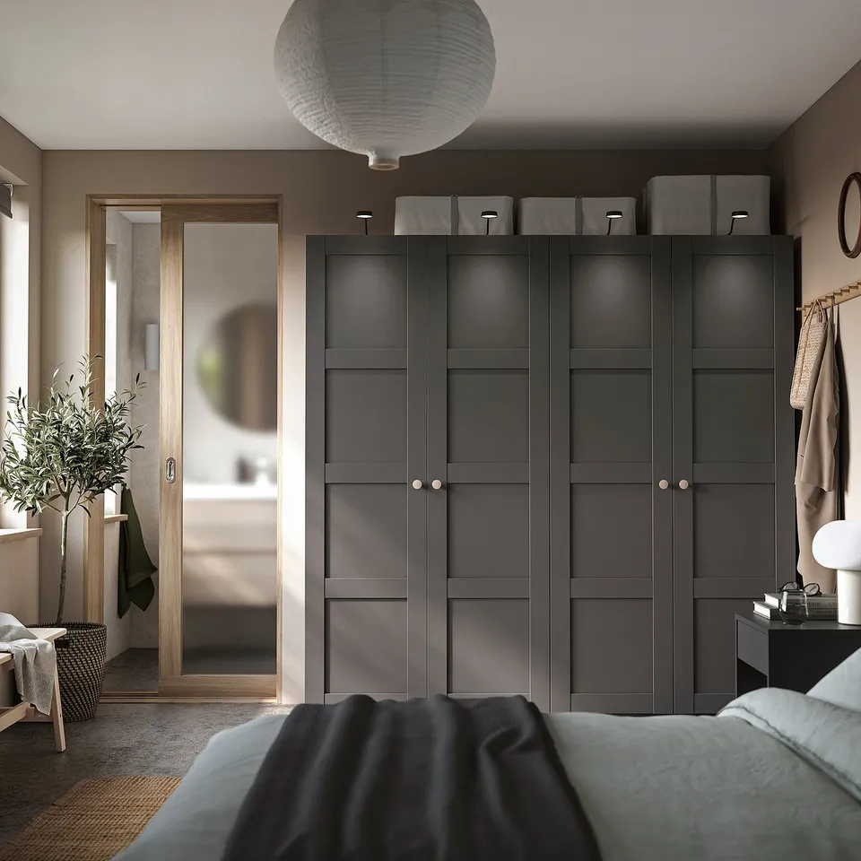 Modern bedroom with dark grey PAX wardrobe, sleek design, natural light, cosy bedding, decorative plants, and a stylish lamp.