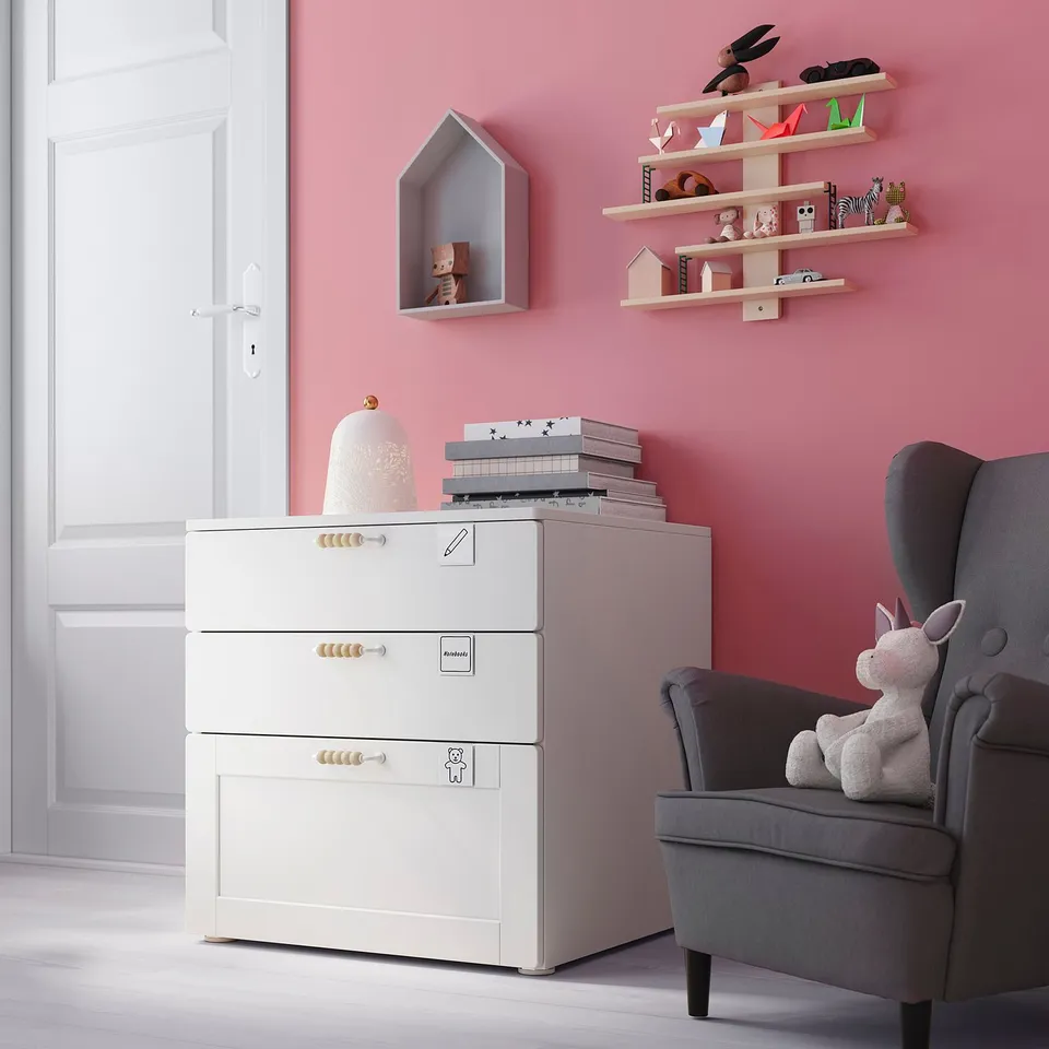 Childs room with pink walls, white storage unit, grey armchair, and shelves with toys.