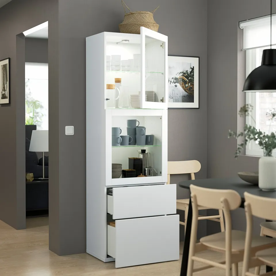 Modern dining area with BESTÅ storage, white cabinet displaying blue mugs and jars, light wood table and chairs.