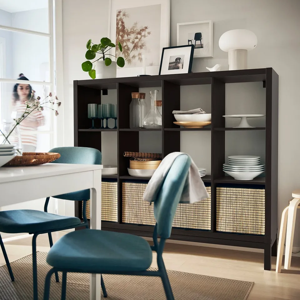 Modern dining area with KALLAX shelf, dishes, decor, blue chairs, person organising on white table. Bright, minimalist decor.