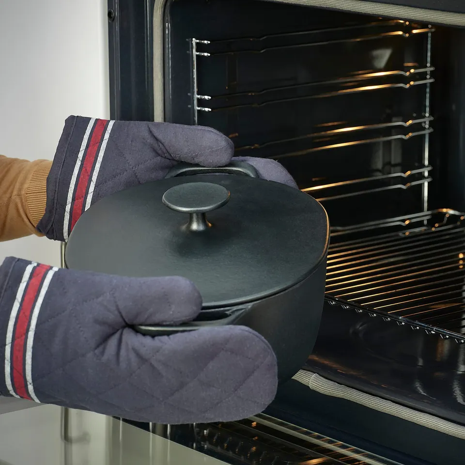 Hands in oven mitts carefully place an enamelled cast iron casserole into an oven, showcasing even heat distribution.