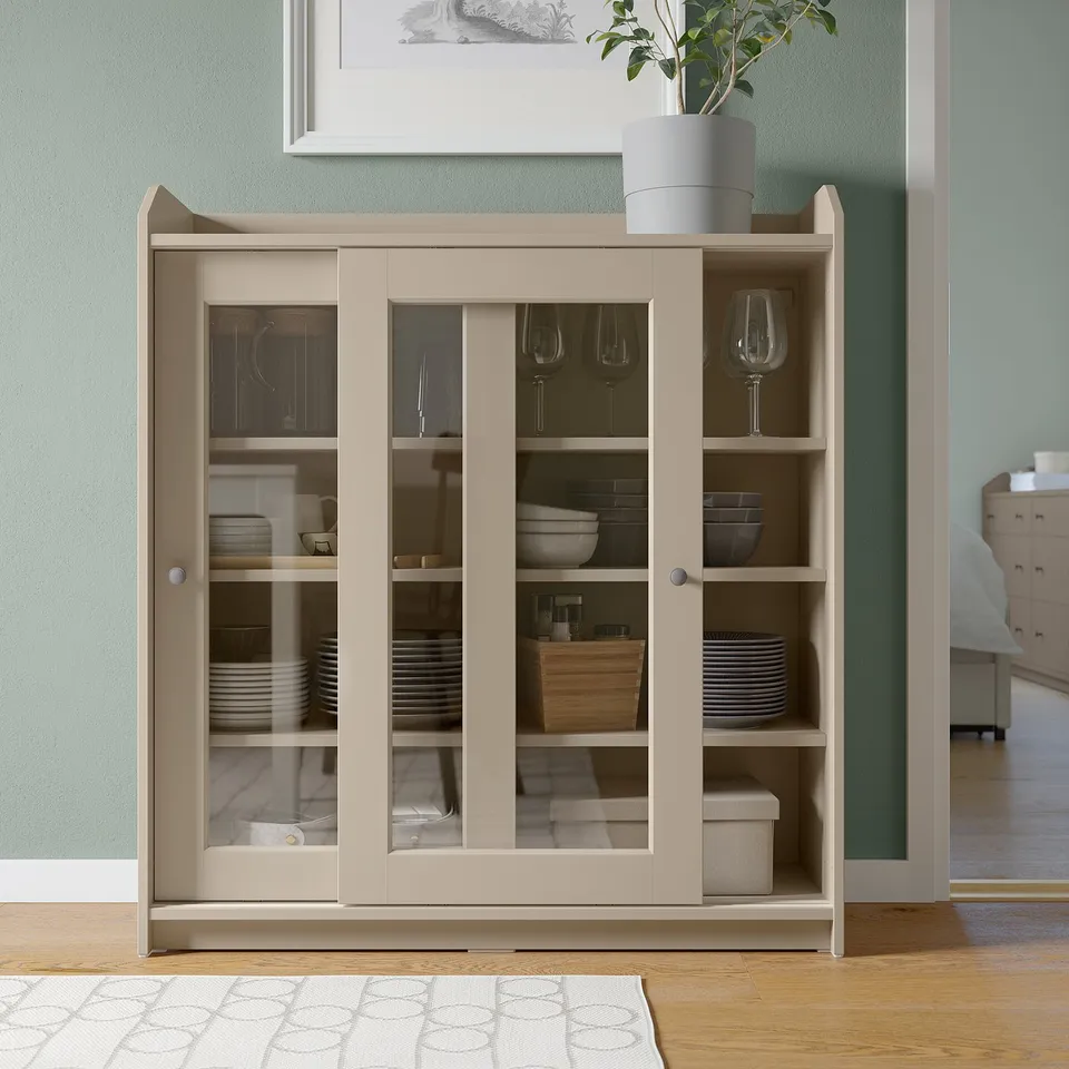Beige HAUGA glass-door cabinet with sliding doors, displaying dishes and wine glasses, positioned against a blue wall.