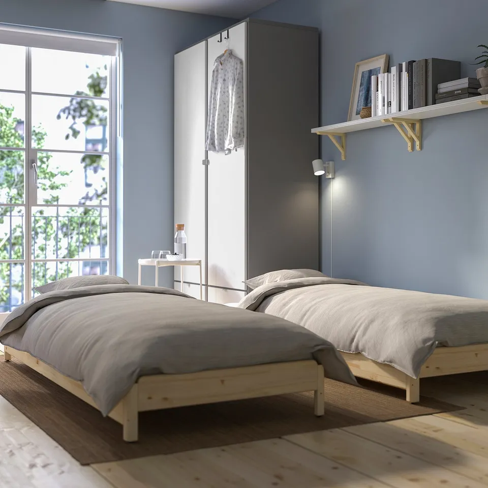 Modern bedroom with two single beds, blue walls, large window, and book shelf. Grey bedding, symmetrical layout.