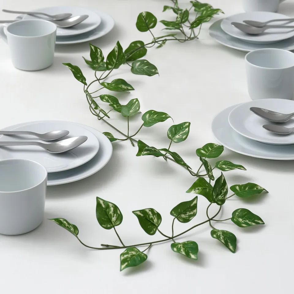 Table set with white plates, cups, and cutlery. A green artificial plant winds between place settings.