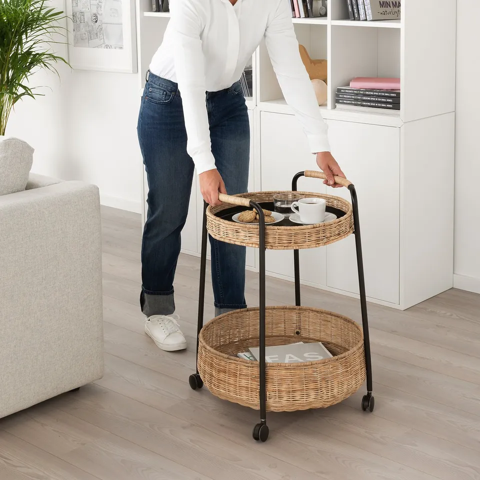 Person pushes woven LUBBAN trolley with coffee, in living room by shelves.