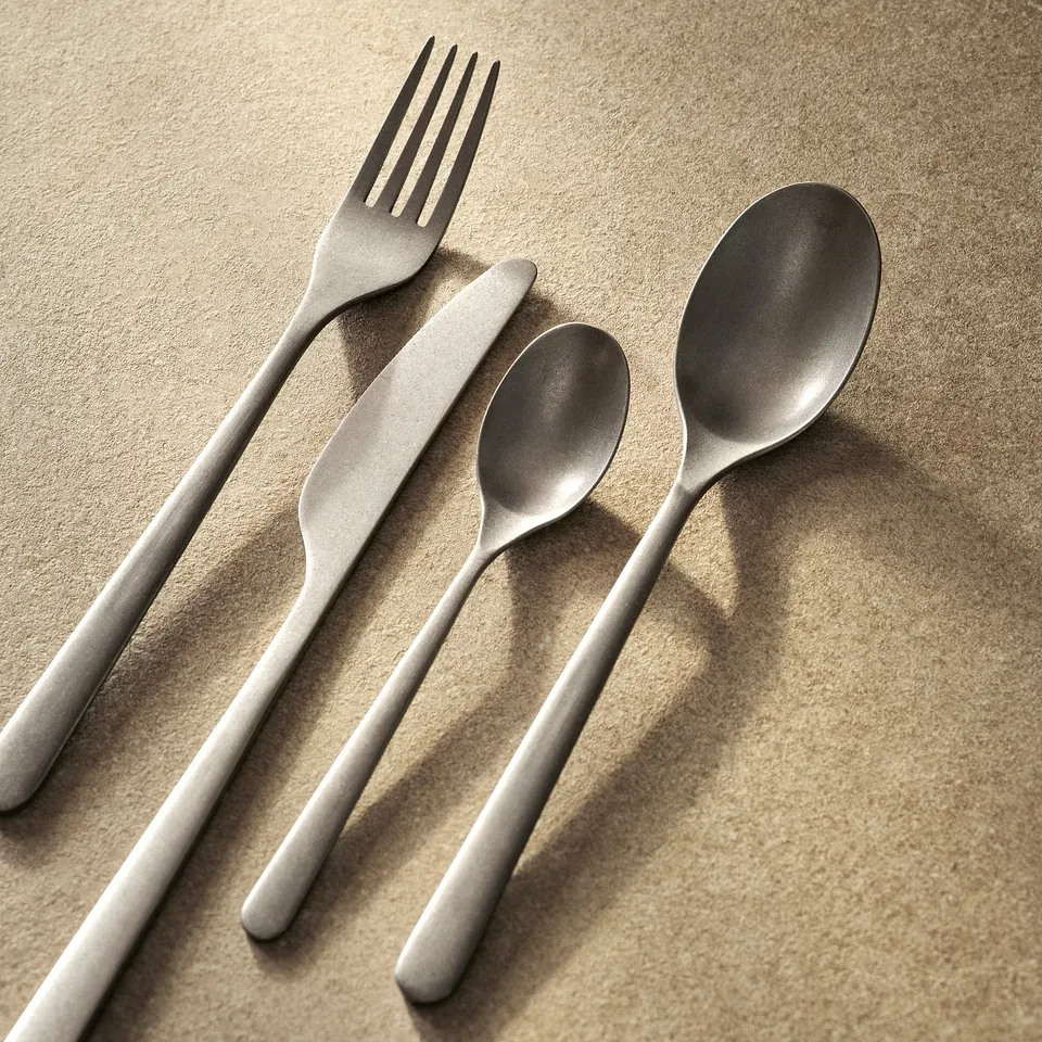 Flat-lay of stainless steel fork, knife, and two spoons with rustic matte finish on beige cloth, showcasing elegant design and quality.