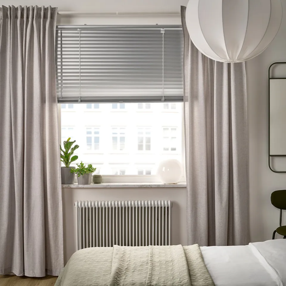 Modern bedroom with VECKLARFLY blinds. Blinds cover window large, light grey, metal slats. Bed below, plants on window sill.