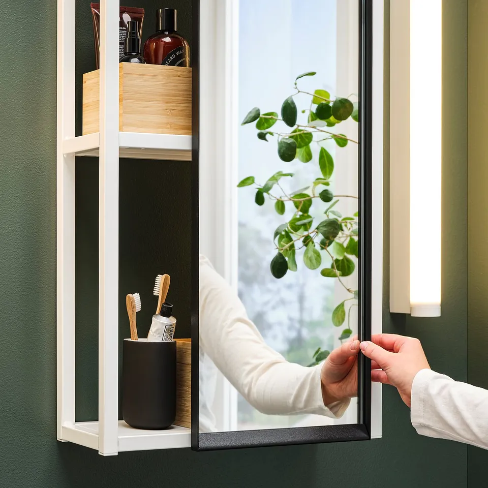 Person opens mirrored cabinet, revealing stored items. Green background, plant reflected.