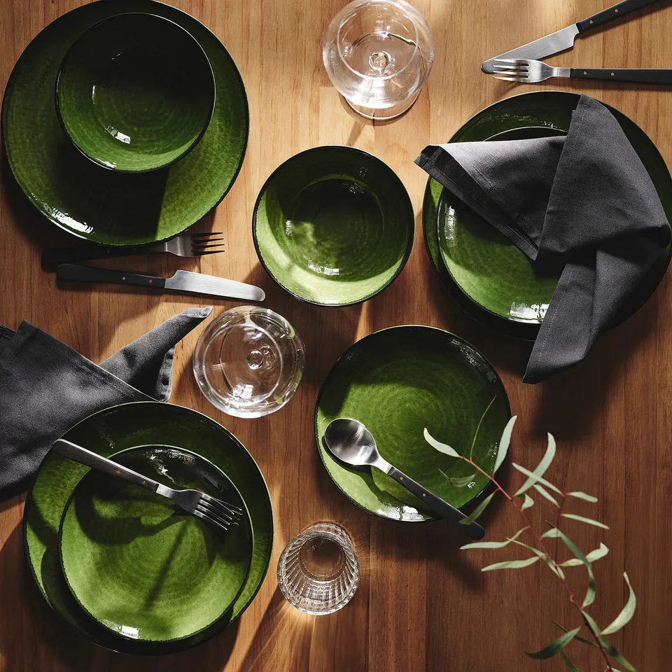 Dark green PELARKAKTUS dinnerware set with glossy finish, including plates, bowls, glasses, and cutlery on wooden table with foliage.