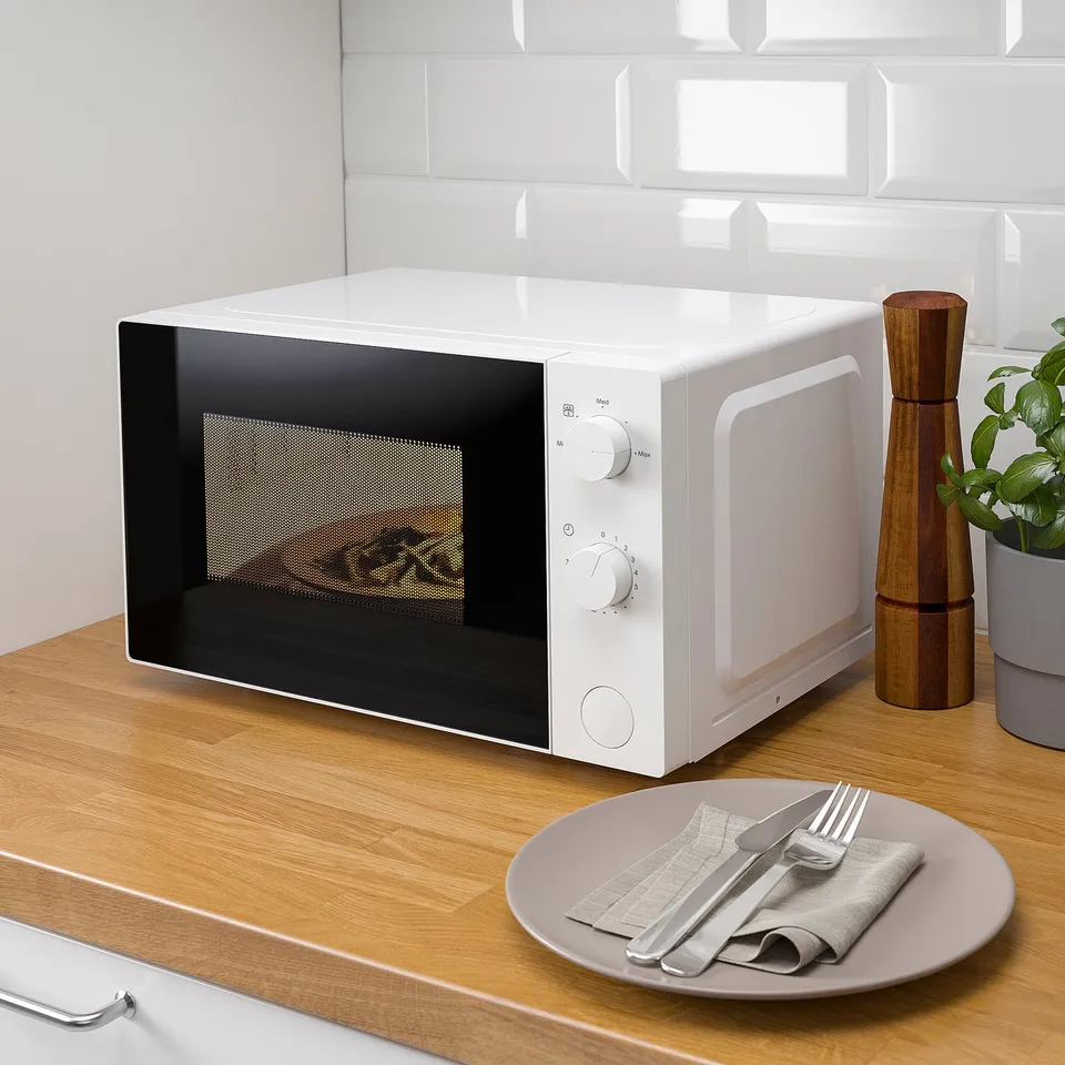 Small kitchen microwave, white, knobs for power and time, silver handles, on wooden counter, beside plate with cutlery.