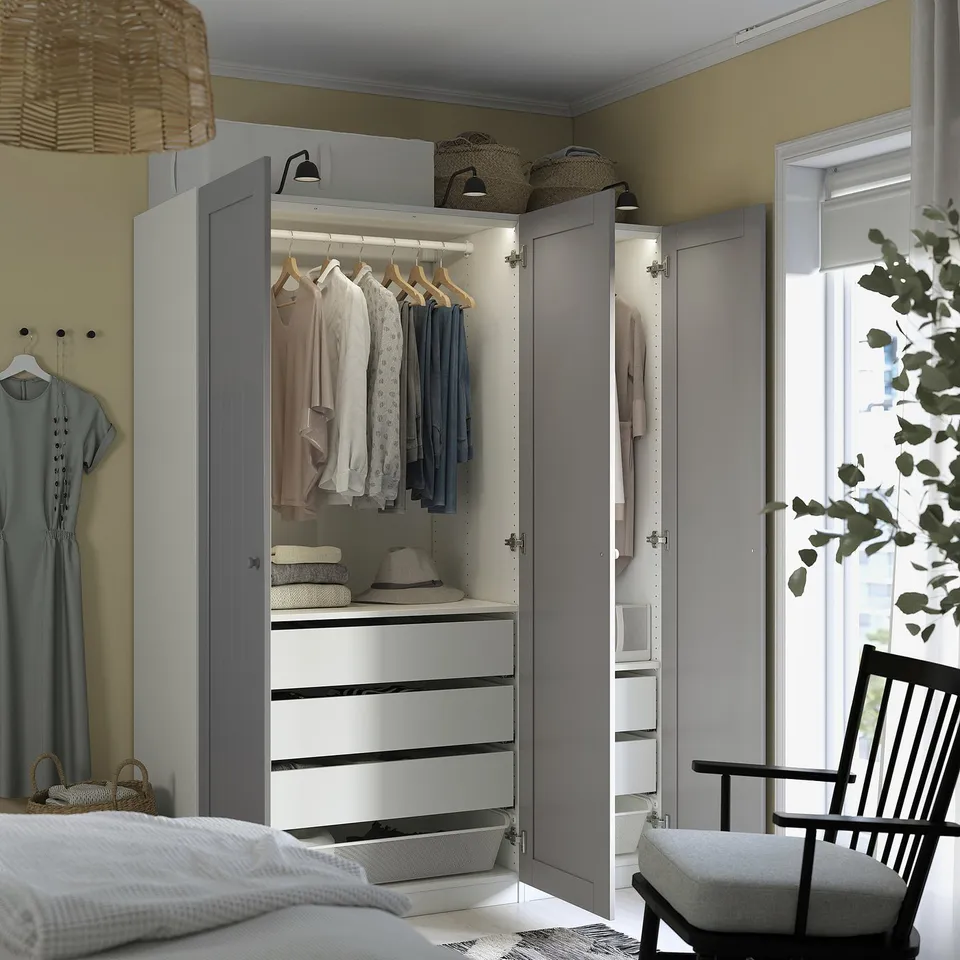 Open wardrobe with clothes, hats, and baskets on top; bed with blanket and chair nearby.