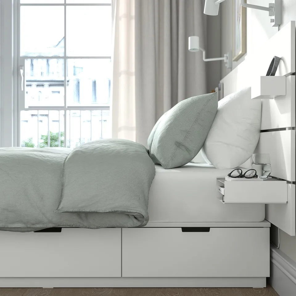 Modern bed with white drawers. Pillows and glasses on bedside table. Soft lighting, city view.