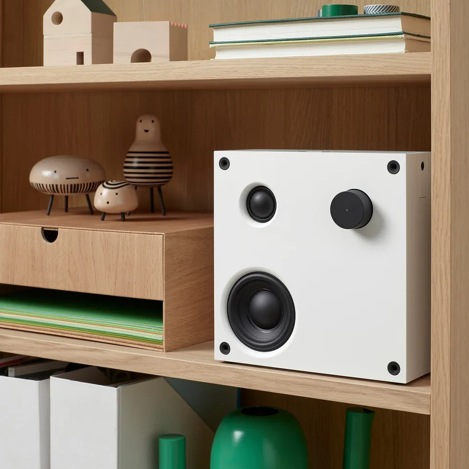 White VAPPEBY bluetooth speaker on wooden shelf, surrounded by decorative items.