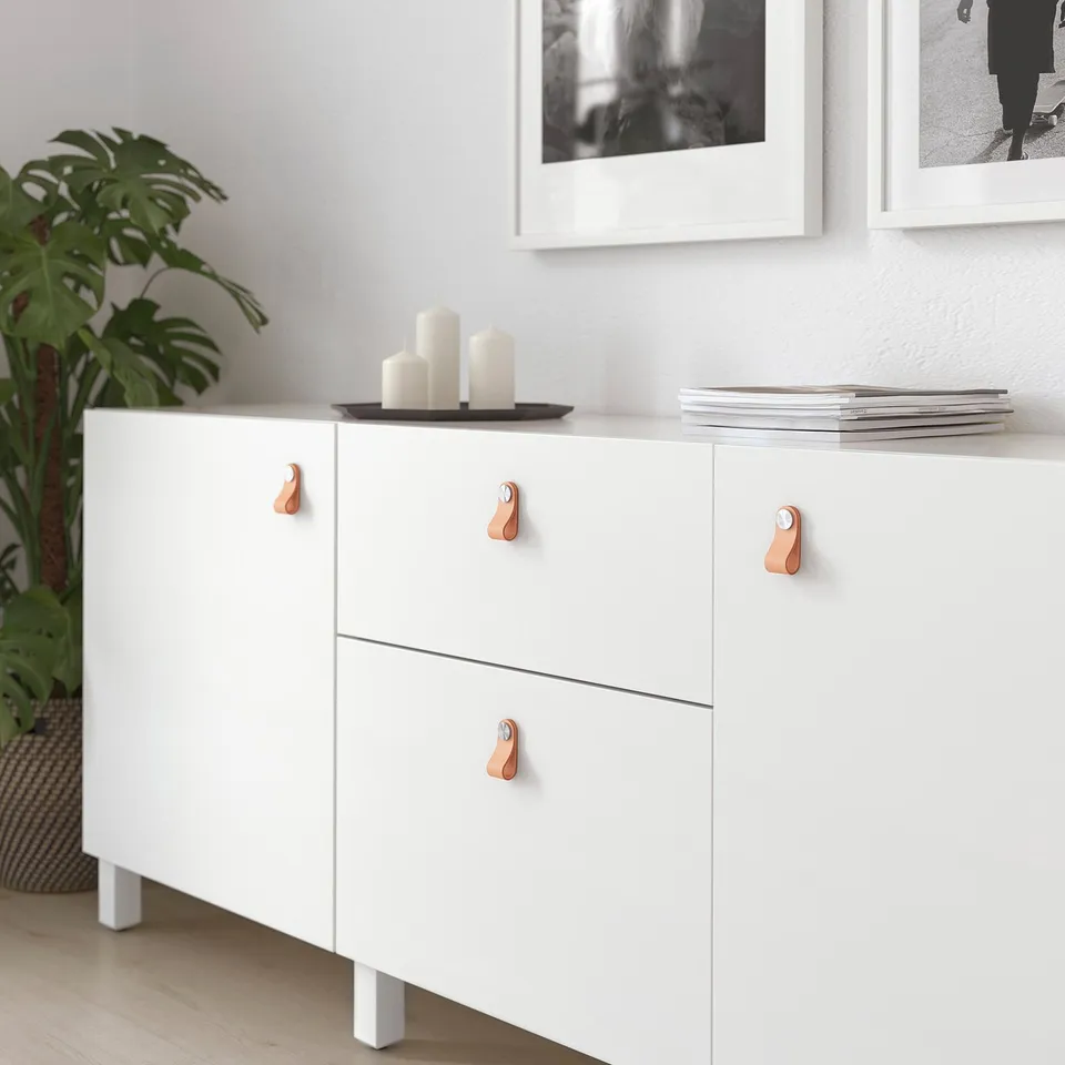 White cabinet with four copper-colored leather ÖSTERNÄS handles; three drawers, two doors. Decorated with candles, tray, and books.