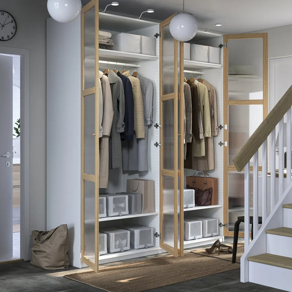 Modern, stylish closet with glass doors holding colourful clothes, and shelves with storage boxes, situated near stairs with hanging lights above.
