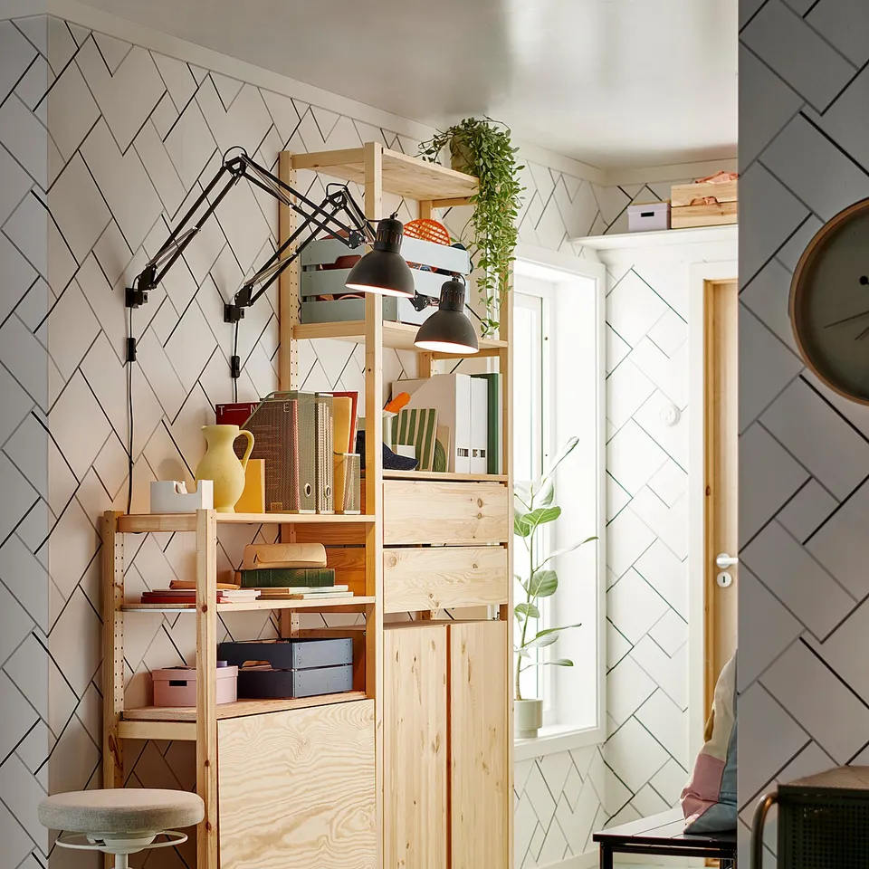 Modern home office with IVAR pine shelving, black lamps, stool, books, and plants. Light-colored wooden floor.