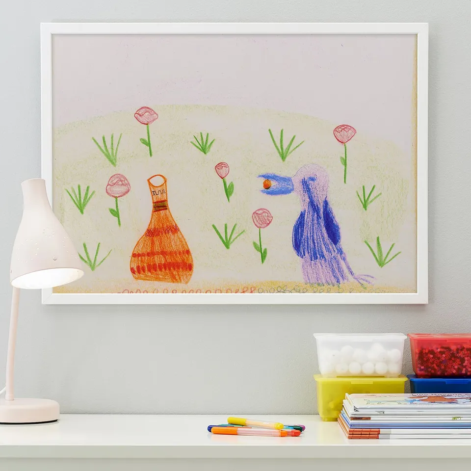 Framed childs drawing with a lamp, markers, and storage bins on a desk.