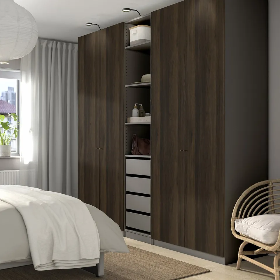 Modern bedroom with large wooden PAX wardrobe, white bed, and wicker chair. Wardrobe is tall, housing shelves and drawers, positioned beside the bed.