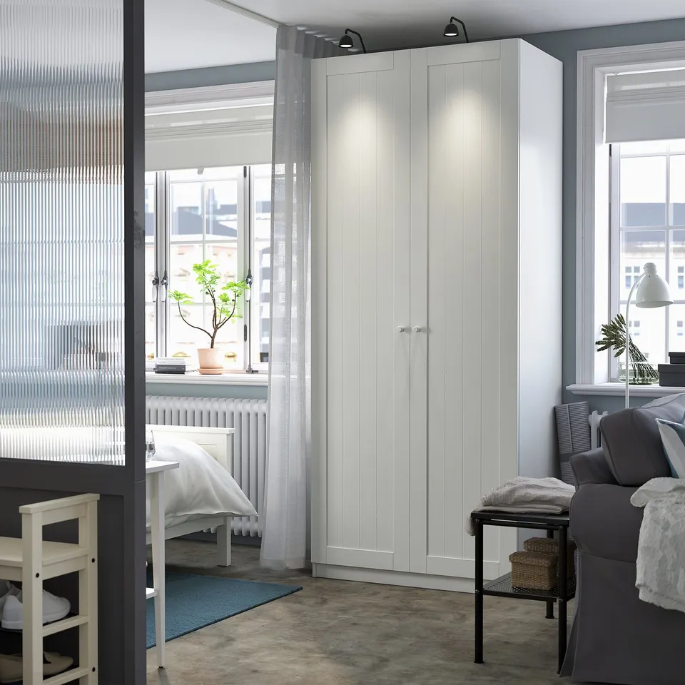 A clean, minimalist room with a prominent white PAX wardrobe, taller than other furniture, featuring sleek panels and a modern design.