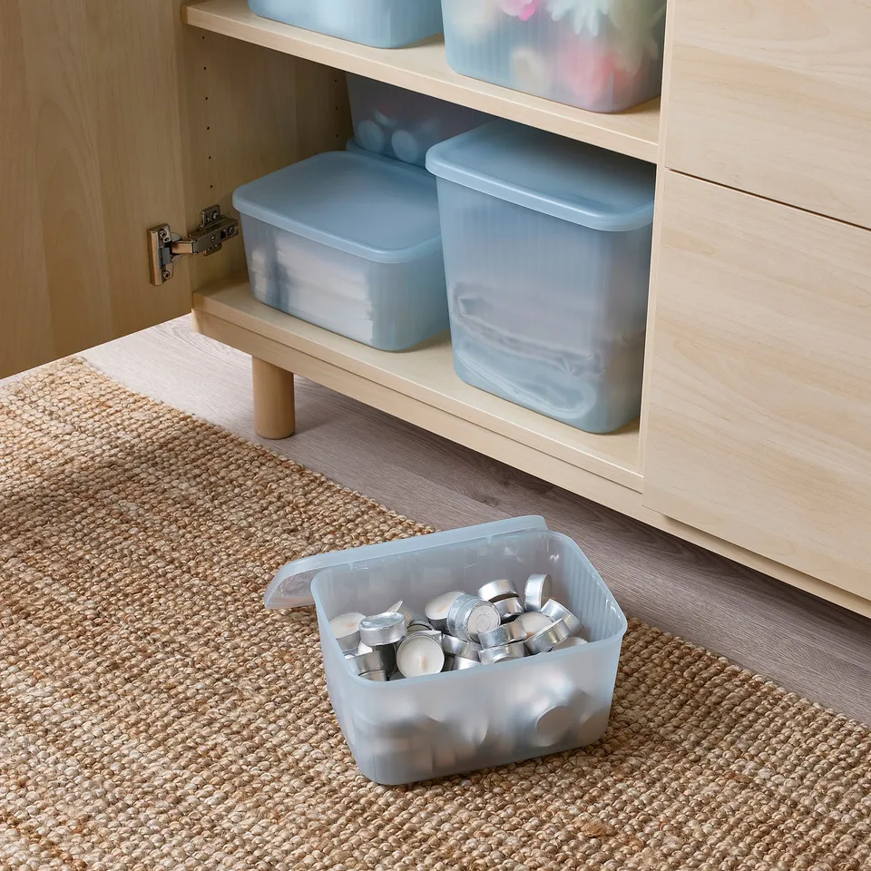 Clear plastic container with lid storing small metal items, sits on woven rug in front of wooden shelves with additional containers.