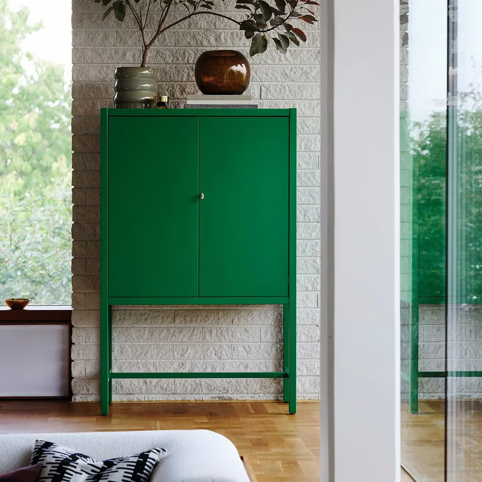 Green STOCKHOLM 2025 cabinet by window, brick wall, glass door, modern decor.
