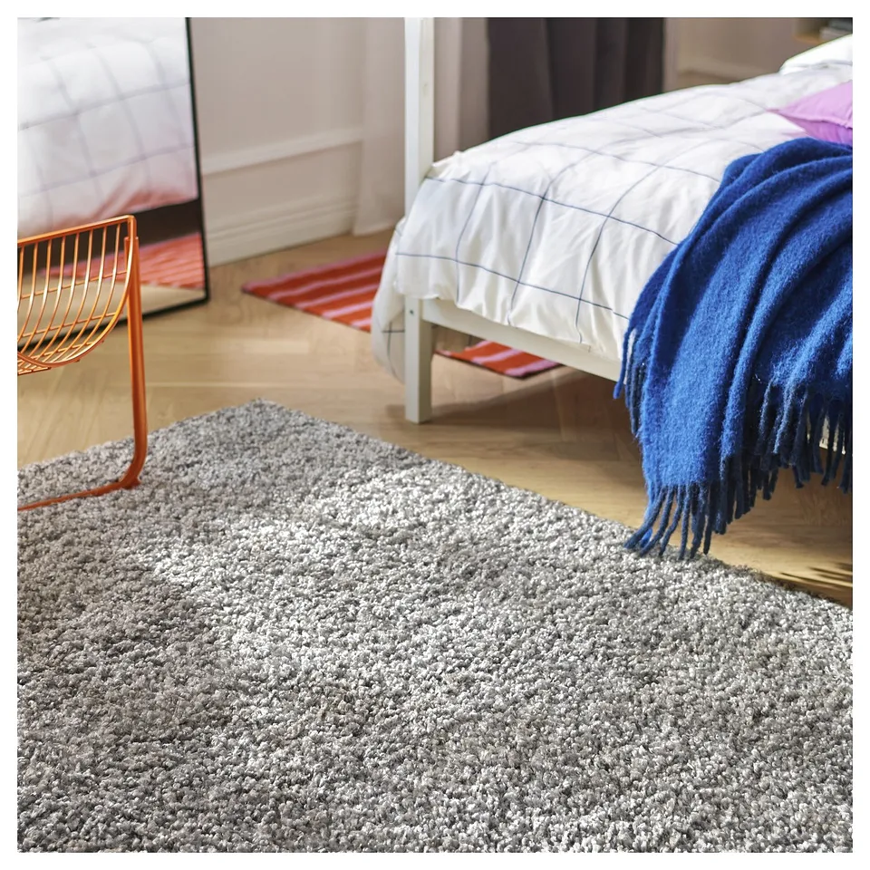 A grey, shaggy ÄRENDE rug in a bedroom with a white bed, blue blanket, and an orange chair.