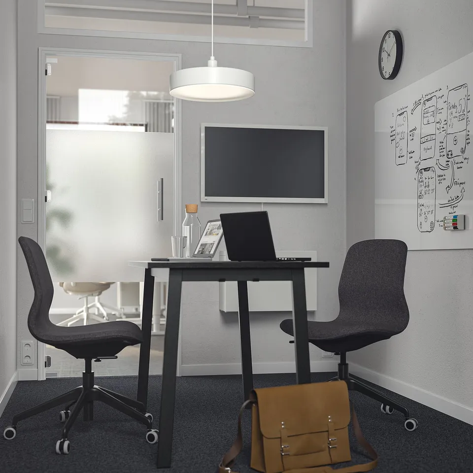 Modern office with MITTZON table, 2 chairs, monitor, laptop, bag, whiteboard, clock, glass door.