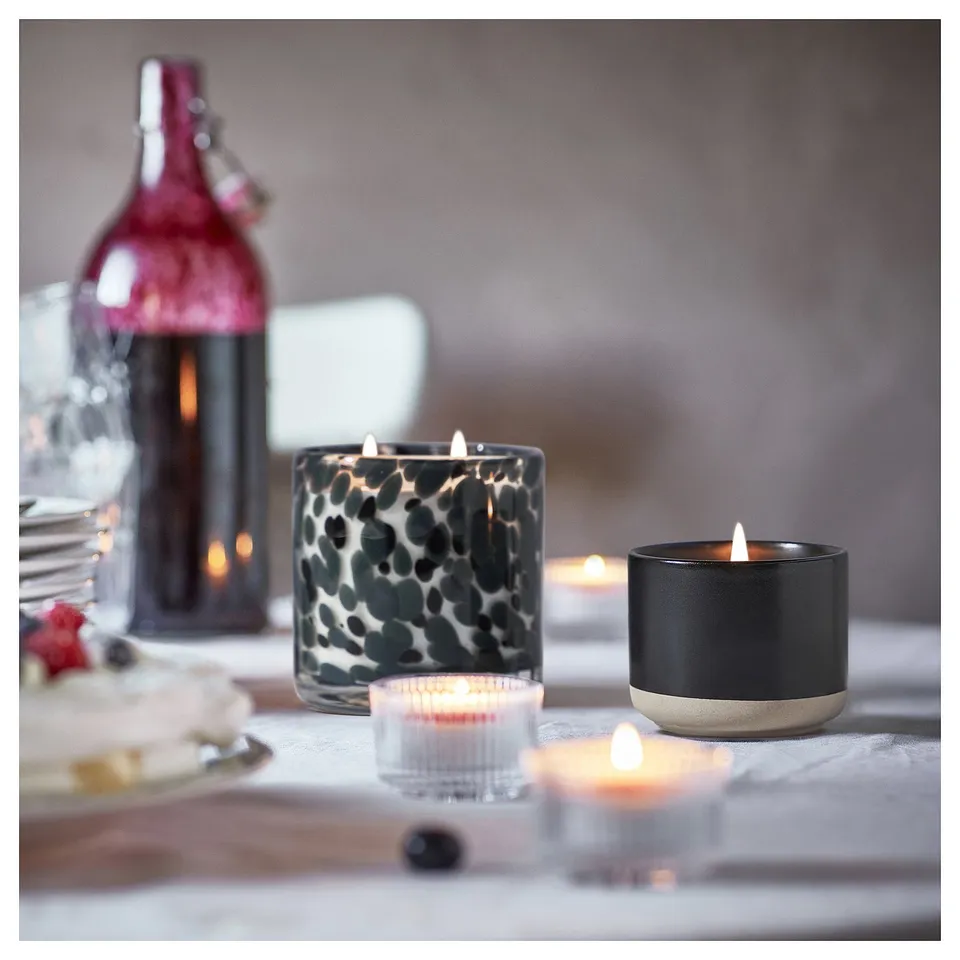 Cosy table setup with lit STÖRTSKÖN candles. Candles in black, patterned, and clear holders. Soft glow on a wooden table.