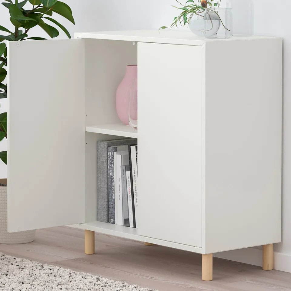 White EKET storage unit with open doors, displaying decorative vase and stacked books. Wooden legs and adjustable shelf.