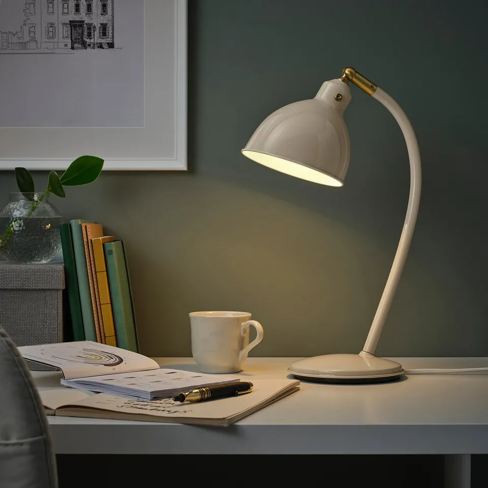 White RÖDFLIK desk lamp with brass on workspace. Includes mug, notebook, pen, books, and wall picture.