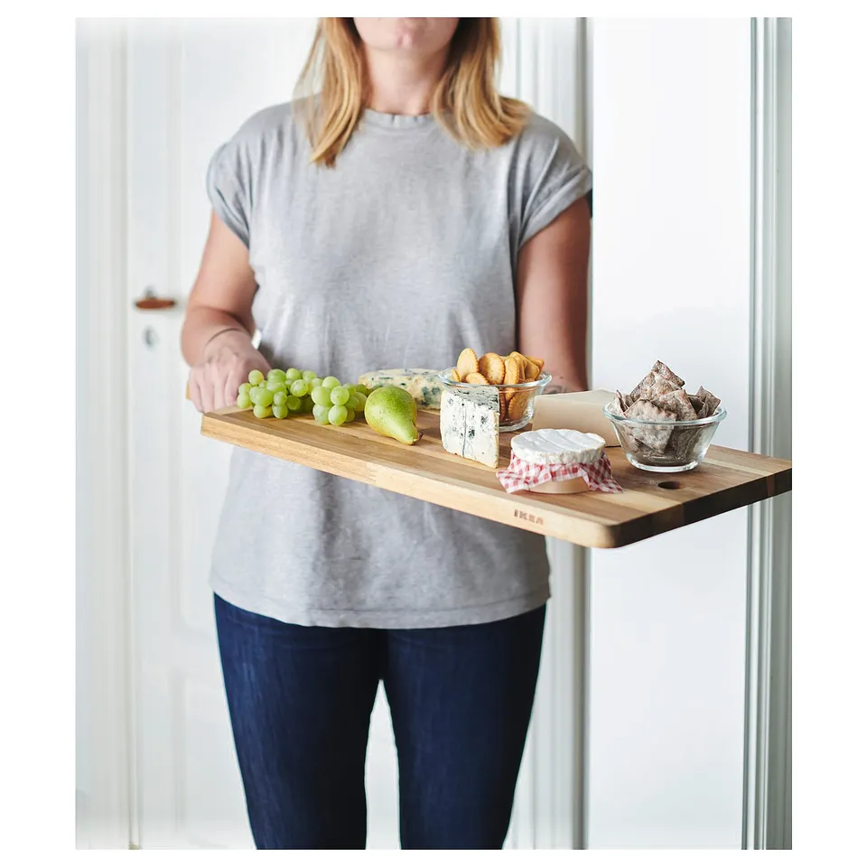 A person holds a wooden serving tray with fruit, cheese, and crackers. The tray is labelled SMÅÄTA.