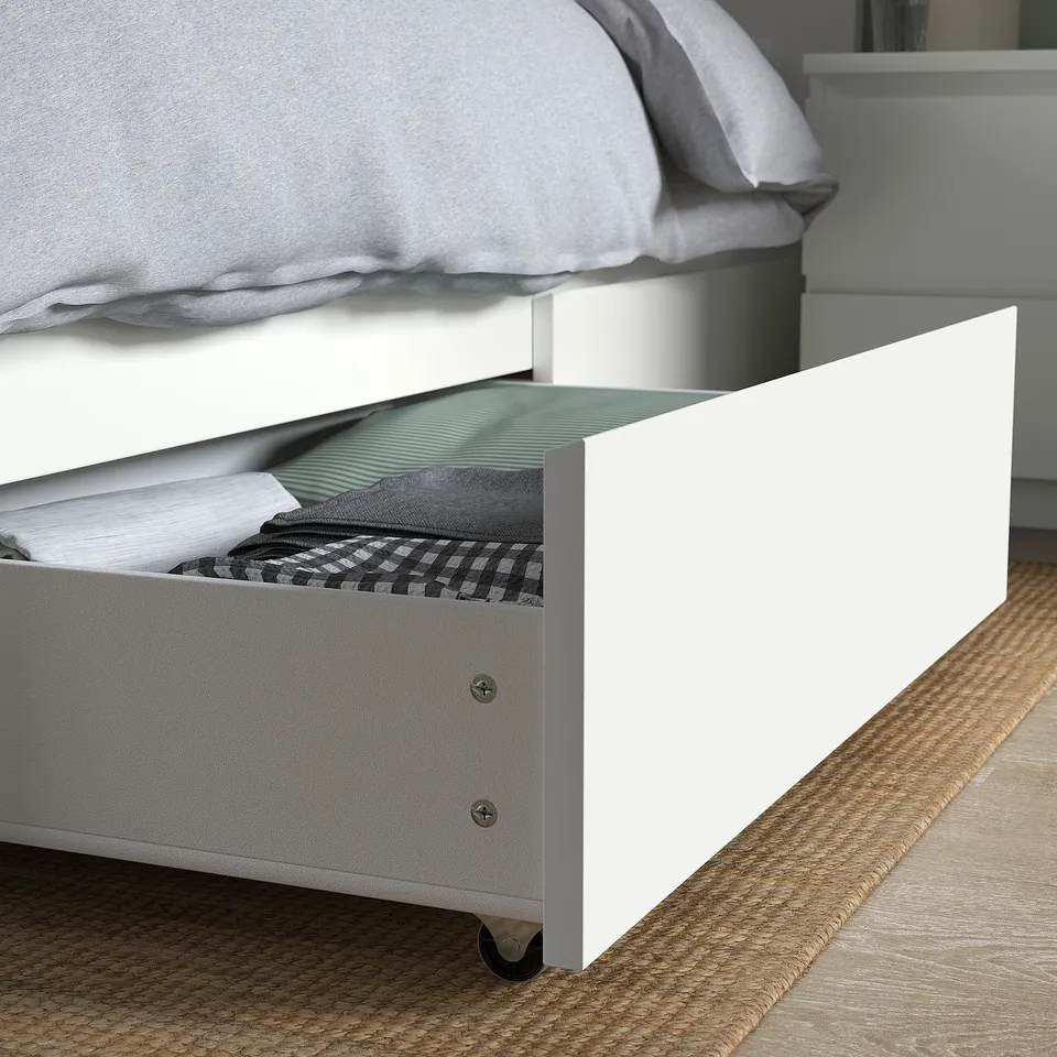 A white MALM bed with a partially open storage drawer revealing folded clothes. The drawer castors for smooth rolling.