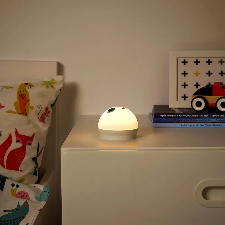 A nightlight named KORNSNÖ on a bedside table, providing a soft glow. Its small and safe, perfect for children.