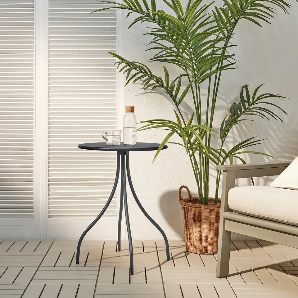 Small anthracite TÅNEBRO side table on patio. Next to large plant, beside white cushioned chair. Metal legs, round top. Lightweight and durable.