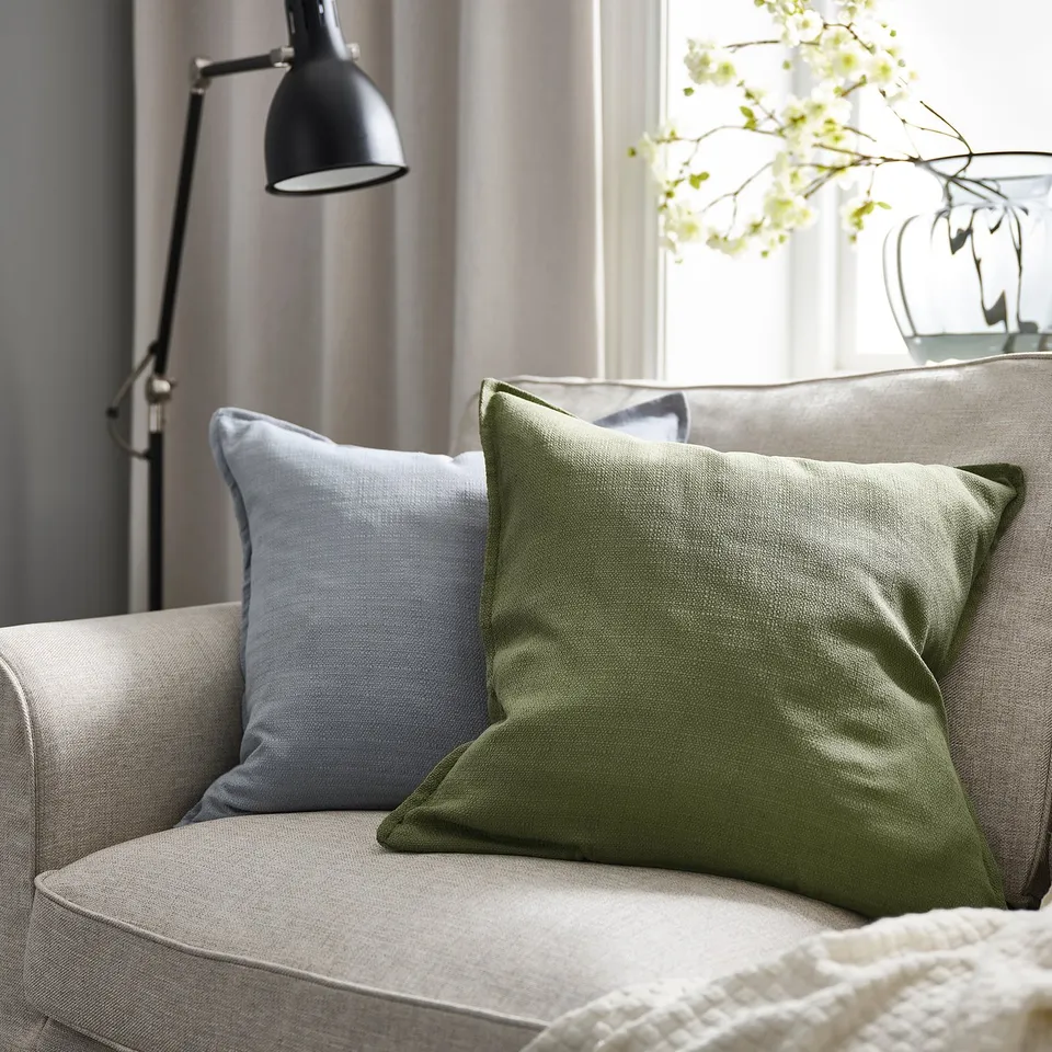 Cosy sofa with grey-green LAGERPOPPEL cushion, textured & zipped, small window with flowers.