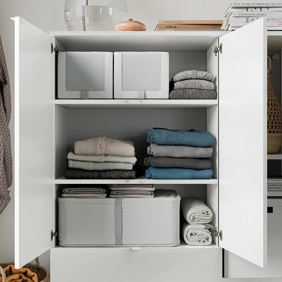 Well-organized white cabinet with neatly folded clothes and storage containers on shelves, topped with decorative items.