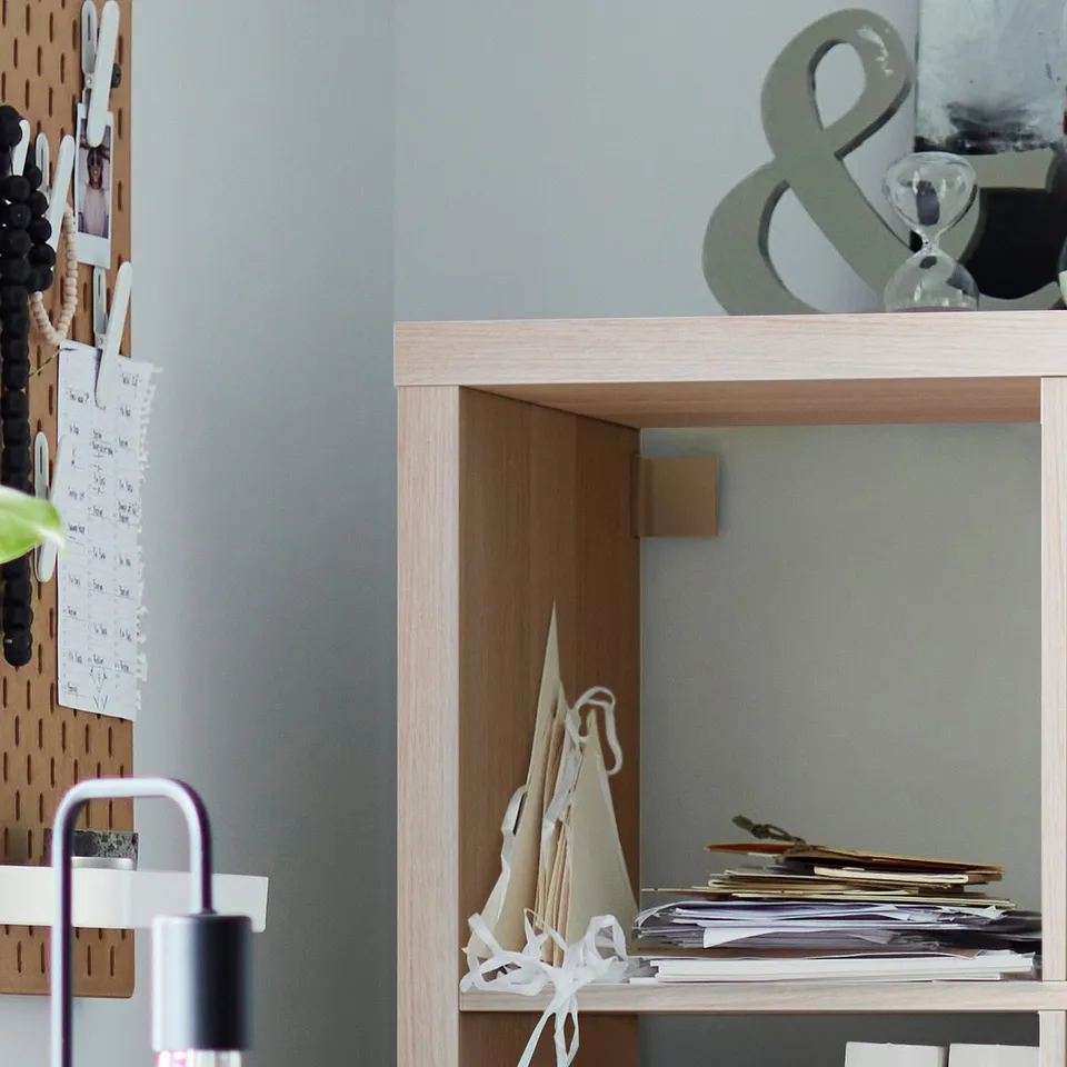 Wooden KALLAX shelving unit with airy underframe, holding papers and cones, against grey wall, next to beige pegboard with notes.
