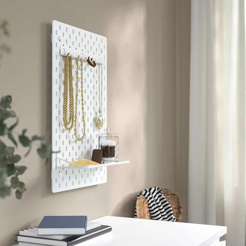 White SKÅDIS pegboard with hooks holding necklaces, shelf with container and wallet, near window with curtains.
