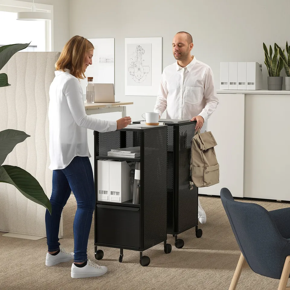 Two people near a BEKANT cart: one hands over papers, showcasing its use as a portable workspace with adjustable shelves.