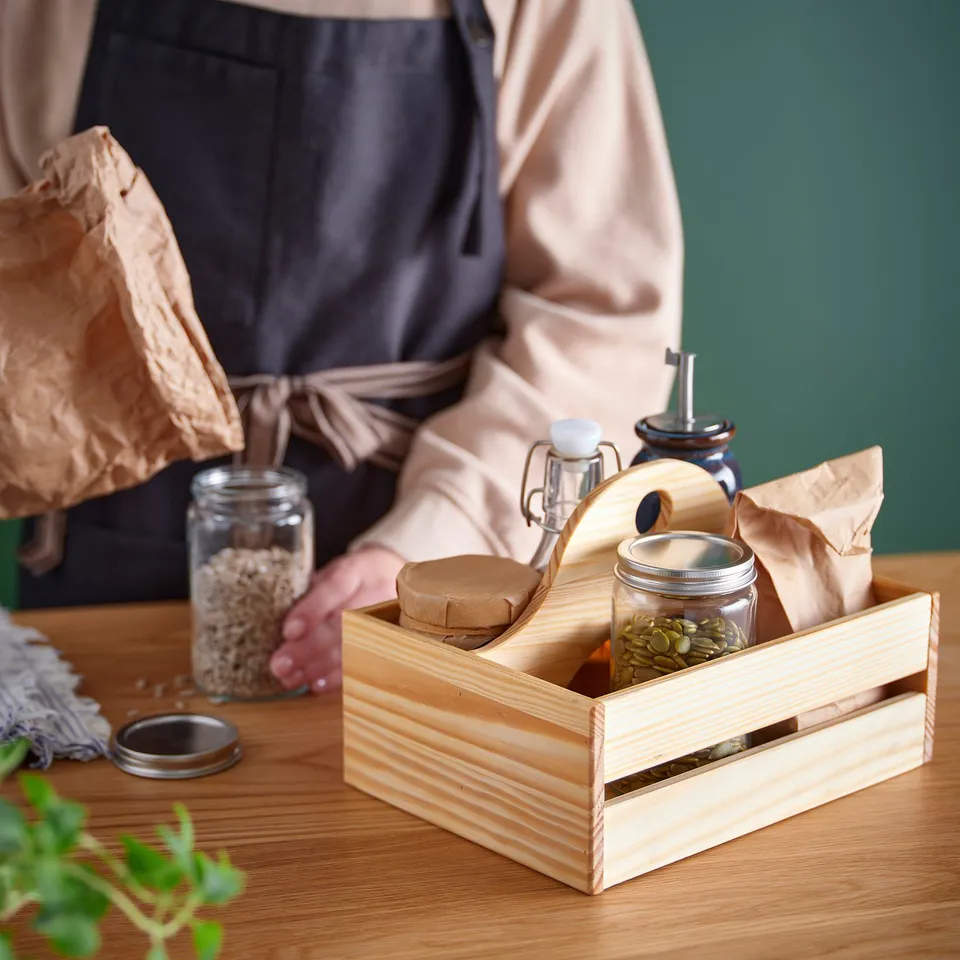 A person holding a wooden crate filled with jars of seeds and bottles, likely related to gardening or plant cultivation.