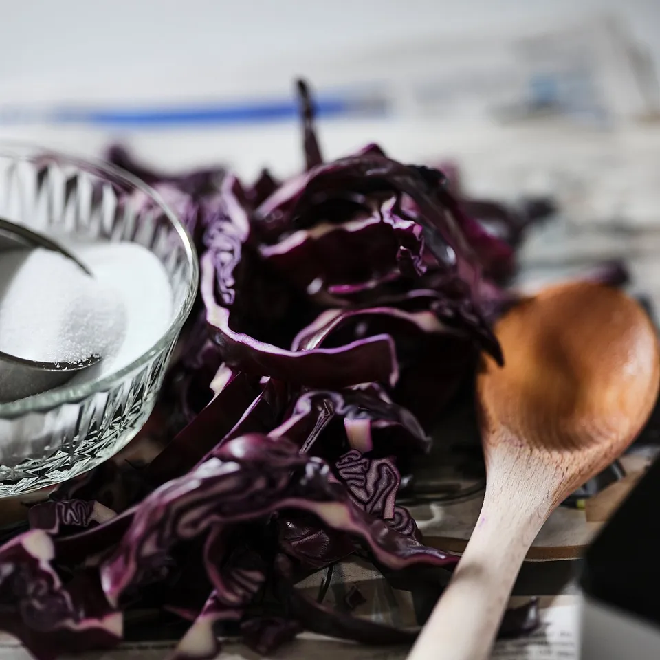 Shredded purple cabbage on a grey counter, next to a wooden spoon, with a container of salt nearby.